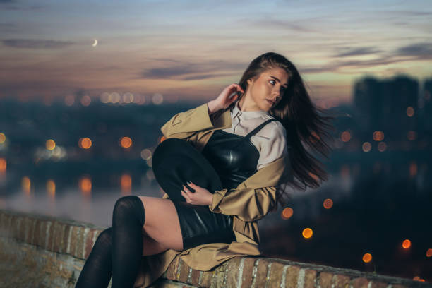 Shot of an attractive young woman sitting on the wall holding the hat in her arms with the dusk lights in the background