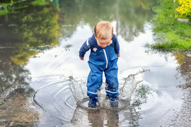 Little cute playful caucasian blond toddler boy enjoy have fun playing jumping in dirty puddle wearing blue waterproof pants and rubber rainboots at home yard street outdoor. Happy childhood concept.