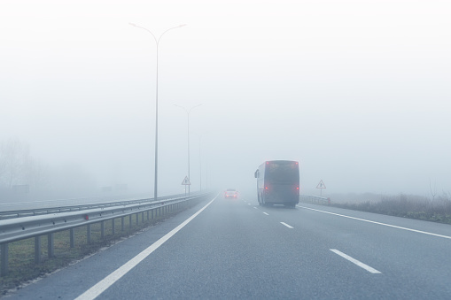 Coach bus on almost empty blue foggy misty rainy highway intercity road with low poor visibility on cold spring autumn morning. Seasonal bad rainy weather accident danger warning. car fog light.