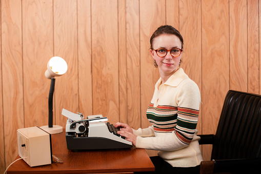 Humor styled photo shoot of a office secretary sitting in a 1970s era desk set up. She has a typewriter on her desk and cheesy wood paneling background.