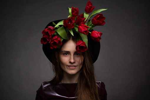 Studio portrait of a young woman in a hat on a black background with a bouquet of red tulips under the hat