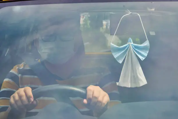 Viewpoint of angel made from protective face mask hanging on rearview mirror of a car while man is driving.