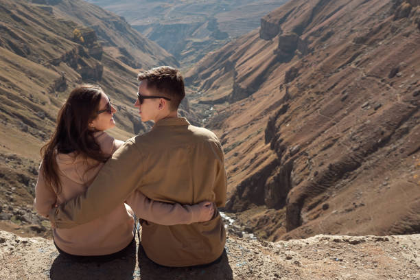 Romantic couple in love hugs and looks into eyes wearing sunglasses and sitting on steep cliff edge against mountain landscape side view