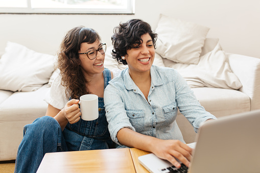 Happy lesbian couple having coffee and using laptop in living room. Beautiful women couple at home with laptop.