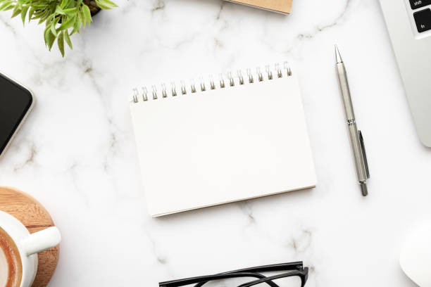 istock Blank notebook with pen are on top of white marble office desk table with laptop computer, cup of coffee and supplies. Top view with copy space. Morning life working from home concept. 1305771444