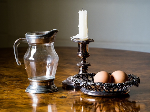 Horizontal closeup of an antique glass and pewter jug, a white candle in an antique silver candlestick and two fresh farm eggs in a decorative silver dish on a rustic wooden table.