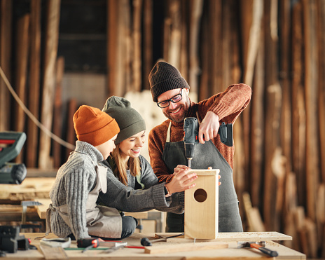 Happy family: focused little boy with mom and dad using drill while making wooden bird house  in professional joinery workshop