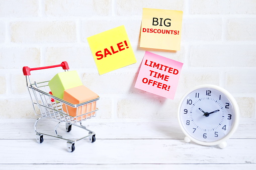 Selective focus of shopping cart or trolley with clock and yellow sticky notes written with 'BIG DISCOUNTS,SALE,LIMITED TIME OFFER'. Shopping theme.