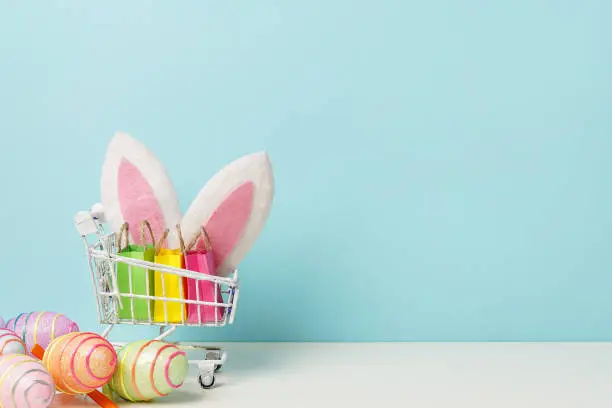 Photo of Colorful painted Easter egg with shopping cart, bunny ears and paper bags , blue background, copy space