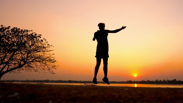 Silhouetted fitness man doing jumping jacks or star jump exercise at seaside outdoors on sunset ,full length portrait.