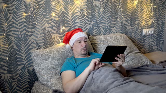 An adult man in a red Santa Claus hat flips through web pages on a tablet while lying on a bed in the bedroom at night. Close-up. The concept of a festive Internet search. 4K.