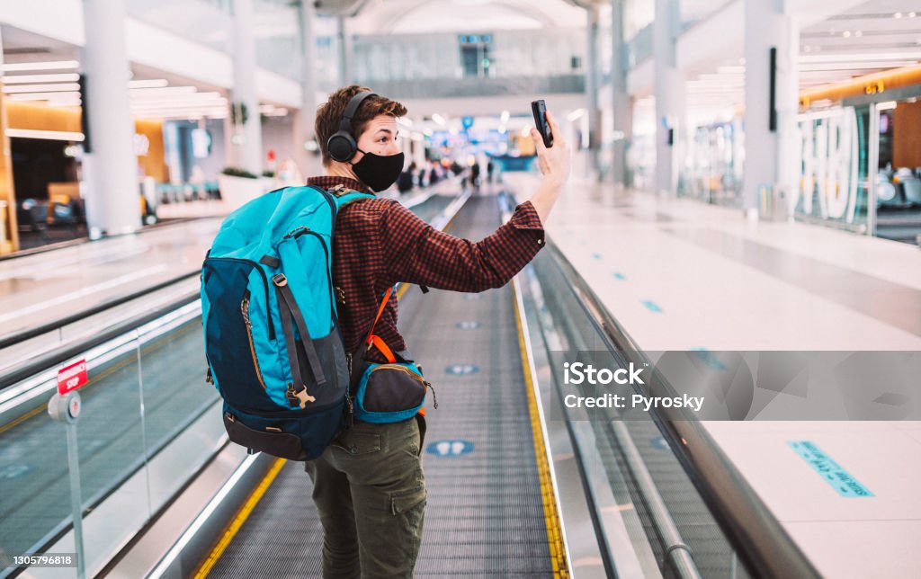 Happy to travel again Rear view of a young man traveler, in a protective face mask, doing selfie, video chatting on a smartphone, while he is standing on an elevated walkway at the airport with a social distancing sign on it.
Traveling during the Coronavirus pandemic Airport Stock Photo