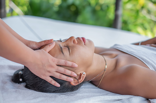 Close-up shot of a beautiful Asian woman lying down on a massage table, having a head massage by an unrecognizable masseuse, in an outdoor spa, close with nature.