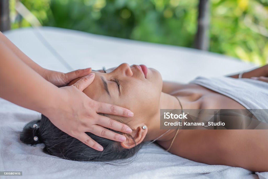 Beautiful Asian Woman Having Head Massage Close-up shot of a beautiful Asian woman lying down on a massage table, having a head massage by an unrecognizable masseuse, in an outdoor spa, close with nature. Massaging Stock Photo
