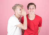 Close-up portrait of two young people, one whispering secrets to the other, shocked and very surprised, with their mouths wide open, isolated on a pink background. Human emotions and facial expressions