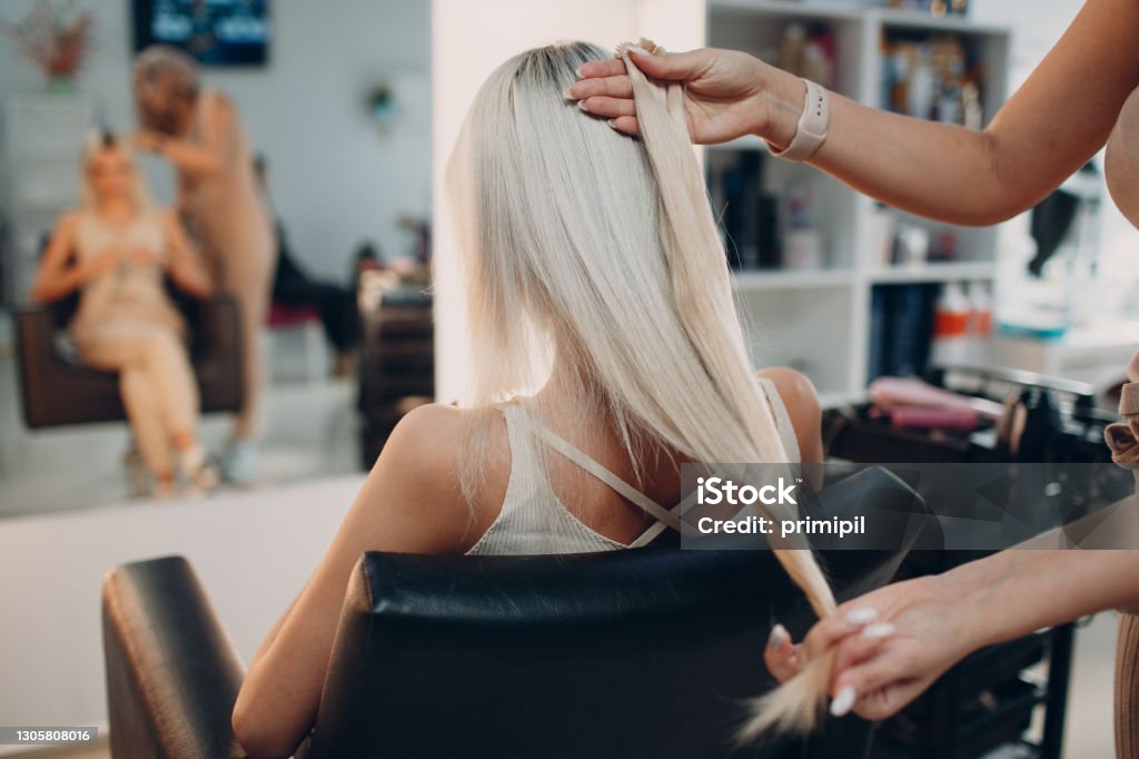 Hairdresser female making hair extensions to young woman with blonde hair in beauty salon. Professional hair extension. Hairdresser female making hair extensions to young woman with blonde hair in beauty salon. Professional hair extension Hair Salon Stock Photo