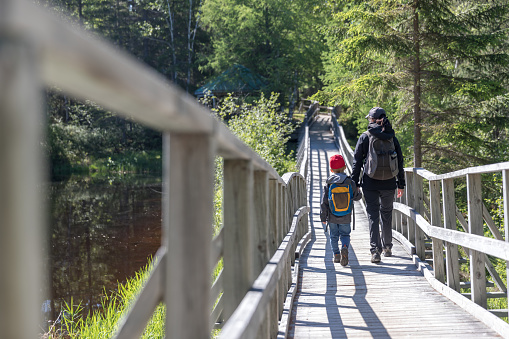 A mother and her preschool age son are crossing a floating wooden bridge over the lake of La Castorière Footpath in Rouyn-Noranda.