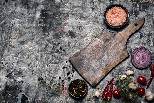 Hand made cutting board on a rustic table with cooking ingredients flat lay tabletop view. An empty slate board is at the center leaving useful copy space for text or logo. Spices and herbs included in the composition