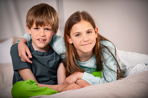Brother and sister in pajamas looking at camera while sitting on bed They are smiling and having fun.