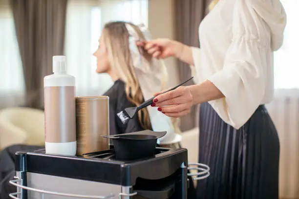 Blonde woman dyeing her hair at the salon. She is sitting and the hairdresser/hairstylist is putting hair color on her hair. This is regular hair care treatment in customers favorite salon.