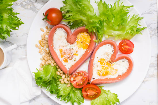 istock Romantic breakfast. Fried eggs in heart shaped sausages, lettuce and cherry tomatoes on a plate on the table. Top view. Close-up 1305837281