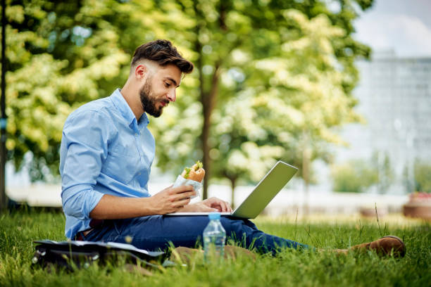 istock Young businessman using laptop eating sandwich in park 1305839322