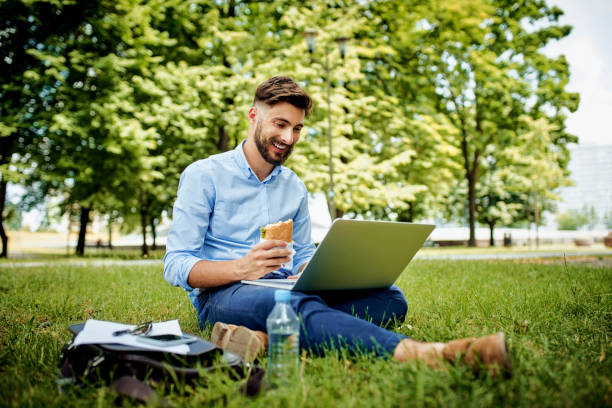 istock Happy young man in park using laptop eating sandwich 1305839442