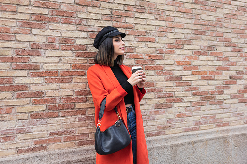 Beautiful young and elegant girl with a coffee. Red coat and black hat. Fashion concept. Brick background.