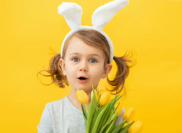 Photo of Easter, holidays, spring concept - happy, funny, crazy, little girl with two ponytails wearing bunny ears headband holding bouquet of yellow tulips and open mouth. Banner, copy space for text, mock up