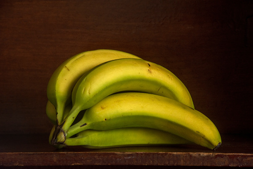 A bunch of unripe bananas are displayed on a wooden shelf in a still life image.