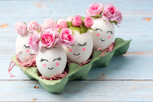 Easter decorative eggs with painted funny cheerful and sleepy faces and floral wreaths in the cardboard egg tray on pastel blue background. Easter creative concept. Copy space, front view.