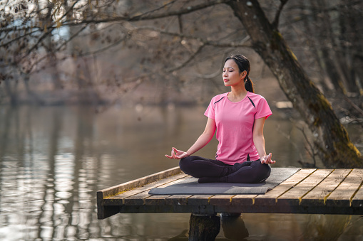 Mid adult Asian woman practicing yoga on the jetty by the lake on a sunny morning. She is meditating.