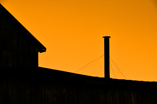A stove pipe exhaust on top of an abandoned barn in front of an orange sky.