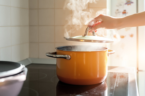 Female hand open lid of enamel steel cooking pan on electric hob with boiling water or soup and scenic vapor steam backlit by warm sunlight at kitchen. Kitchenware utensil and tool at home background.