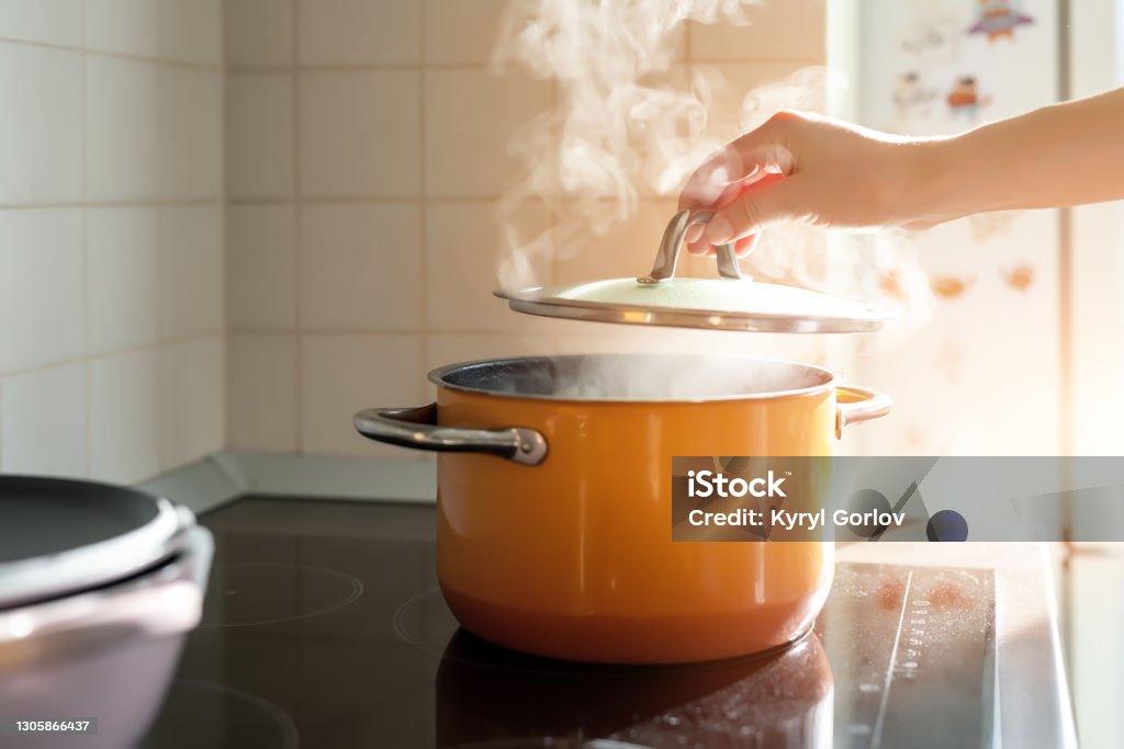 Female hand open lid of enamel steel cooking pan on electric hob with boiling water or soup and scenic vapor steam backlit by warm sunlight at kitchen. Kitchenware utensil and tool at home background Female hand open lid of enamel steel cooking pan on electric hob with boiling water or soup and scenic vapor steam backlit by warm sunlight at kitchen. Kitchenware utensil and tool at home background. Cooking Pan Stock Photo