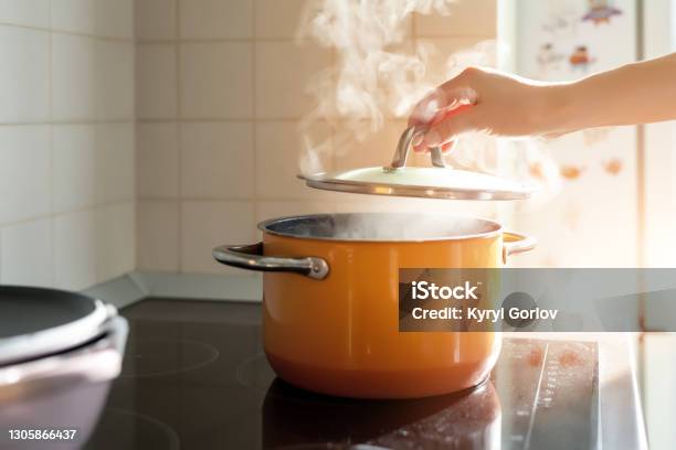 Female Hand Open Lid Of Enamel Steel Cooking Pan On Electric Hob With Boiling Water Or Soup And Scenic Vapor Steam Backlit By Warm Sunlight At Kitchen Kitchenware Utensil And Tool At Home Background Stock Photo - Download Image Now