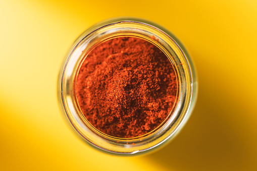 Powdered red pepper in glass jar on yellow background. Spices from directly above, selective focus.