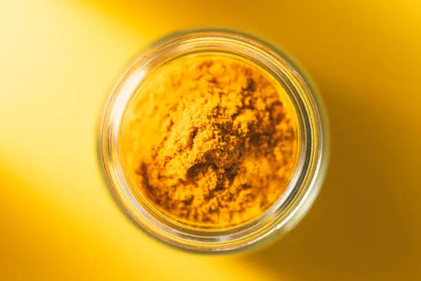 Turmeric powder in glass jar on yellow background. Spices from directly above, selective focus.