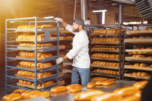 пекарь несет стеллаж с хлебом в пекарне. промышленное производство хлеба - пекарь стоковые фото и изображения