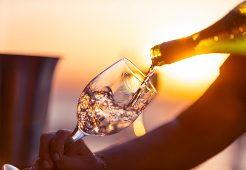 Close up of white wine being poured into a glass.  A second glass can be seen in the background. A woman is holding the glass and pouring the wine. Ice bucket and wine bottle with a romantic sunset.