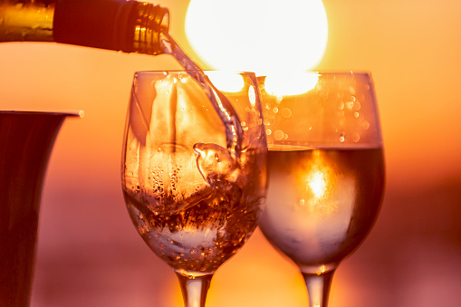 Close up of white wine being poured into 2 glasses. Ice bucket and wine bottle with a romantic sunset. Glass is empty