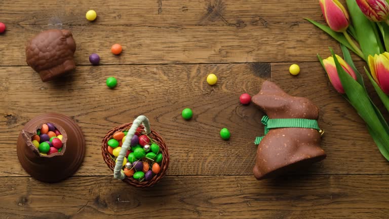 Top down view on delicious chocolate Easter bunny and colorful candy in a basket next to bouquet with tulips on wooden table. A hand puts a chocolate rabbit on the table.