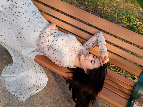 Top view of beautiful Chinese young woman in white dress lying on park bench in sunny day.