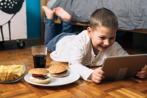 boy eating junk food and using digital tablet or laptop computer at home little boy digital tablet and eating junk food in his room terrible meal stock pictures, royalty-free photos & images