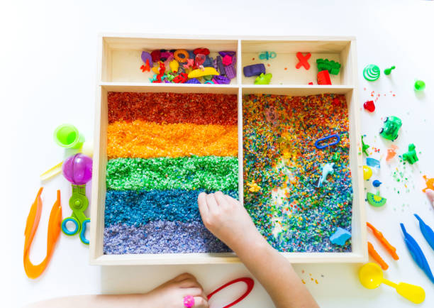 Sensory box with rainbow rice inside. Children's hands touch the texture and pour. Tool for sorting tongs and spoon. Montessori material baby