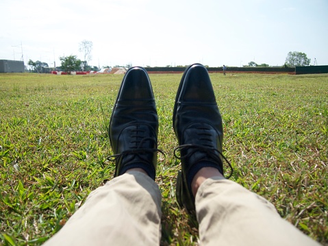 The legs of a man wearing leather shoes sitting on the grass.