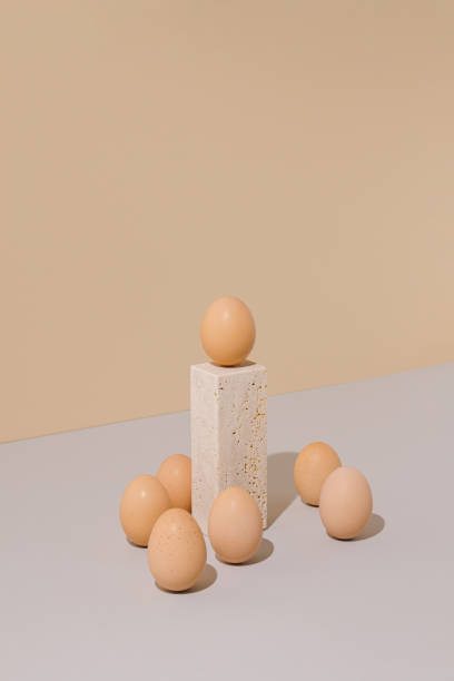 istock Single egg standing on a travertine marble block podium surrounded by group of eggs on a beige and gray background. 2021 Easter unique still life composition. Surreal retro creative layout. 1305917979