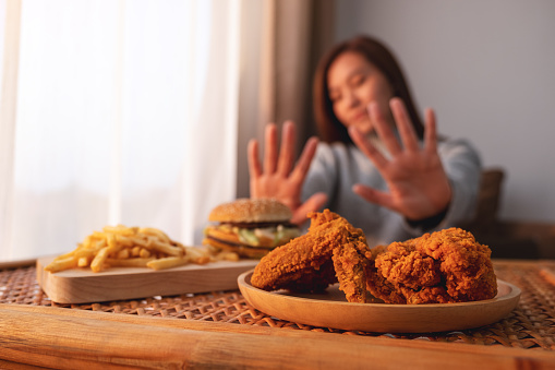 A woman making hand sign to refuse a hamburger, french fries and fried chicken on the table for dieting and healthy eating concept