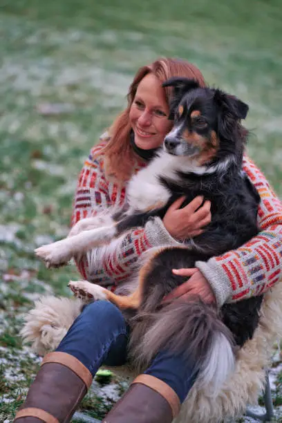 Blonde woman with her Australian Shepherd dog on her lap. In casual clothing, winterboots and thick winter sweater Outside on the snowy grass by the campfire in winter.