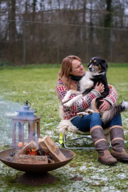 Blonde woman with her Australian Shepherd dog on her lap. In casual clothing, winterboots and thick winter sweater Outside on the snowy grass by the campfire in winter.